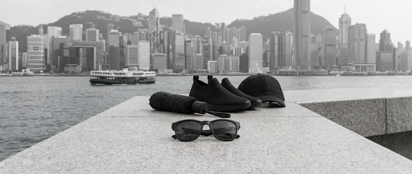 Black cap, sunglasses, compact umbrella and slip-on sneakers with the Hong Kong skyline behind