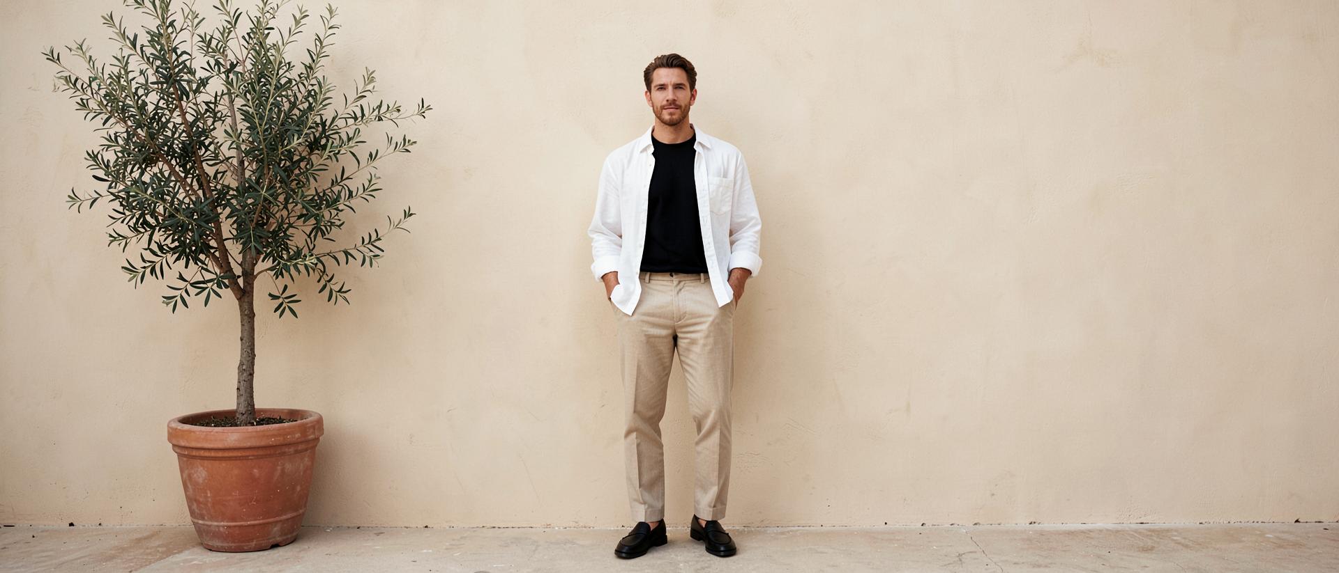 A man wearing a white linen shirt open over a black T-shirt with stone trousers and black loafers